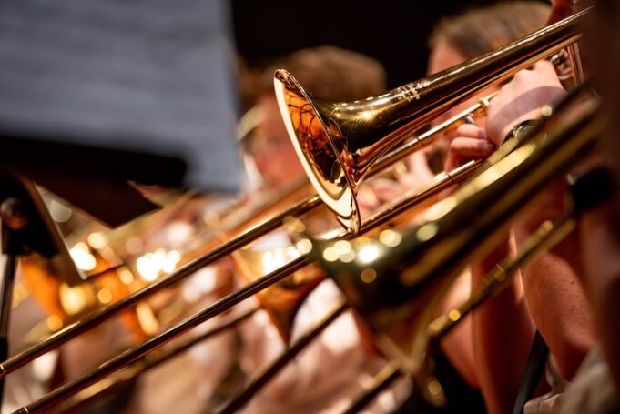 Close-up view of high school band students performing with brass instruments, including trumpets and trombones, during a school concert. The image captures the golden metallic shine of the instruments