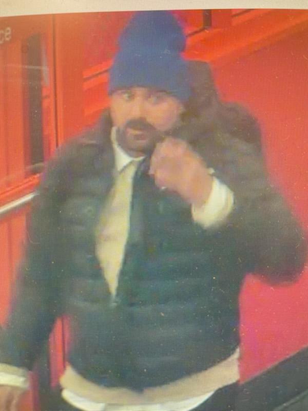 A surveillance-style photo of a man indoors wearing a blue beanie and dark jacket, with a red/orange background. No readable text is visible in the image.