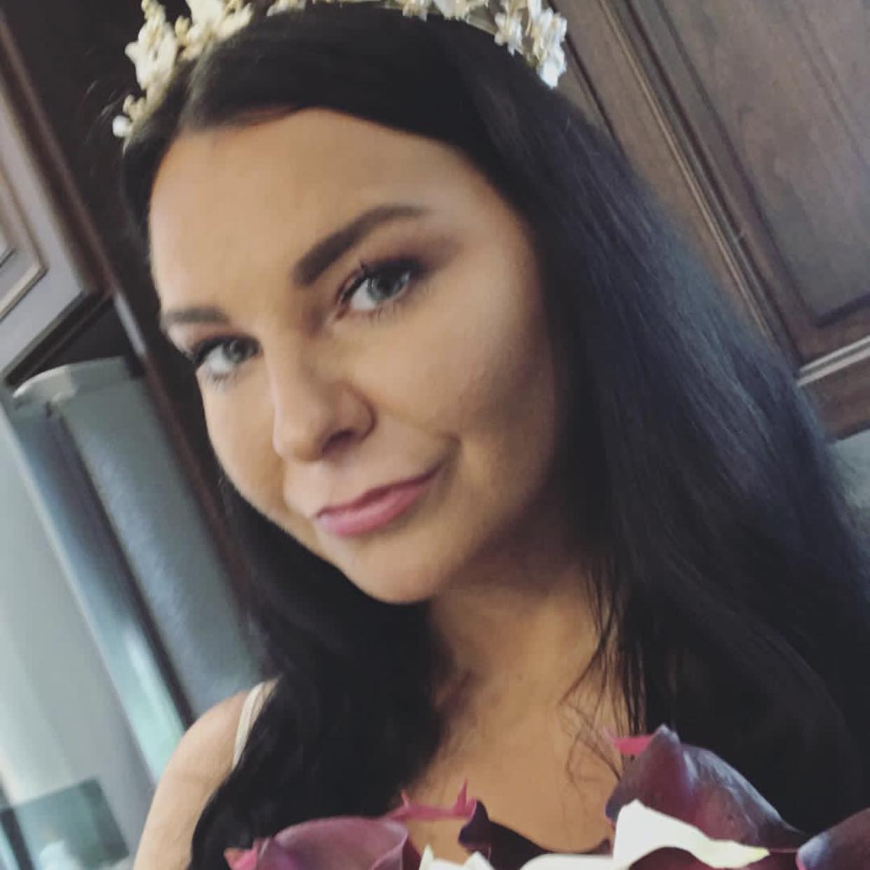 A close-up portrait photo of a woman with dark hair wearing a floral headpiece, indoors. No visible text appears in the image.