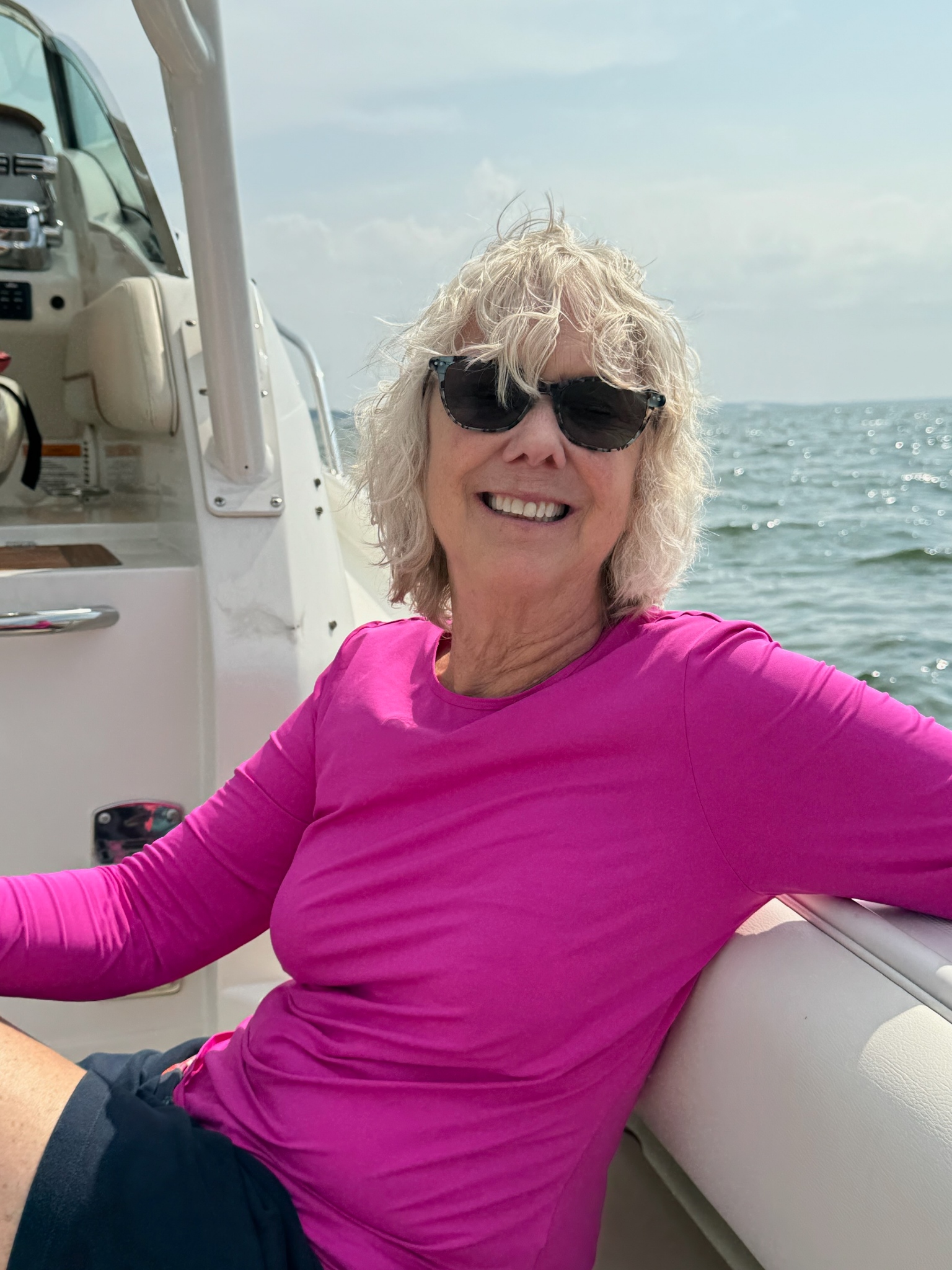 A candid photo of a woman wearing sunglasses and a bright pink top sitting on a boat, with the ocean and sky in the background. No readable text is visible.