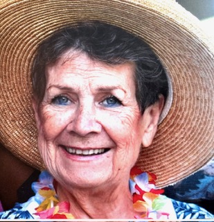 A close-up portrait photo of an older woman smiling, wearing a wide-brimmed straw hat and a colorful flower lei. No readable text is visible in the image.