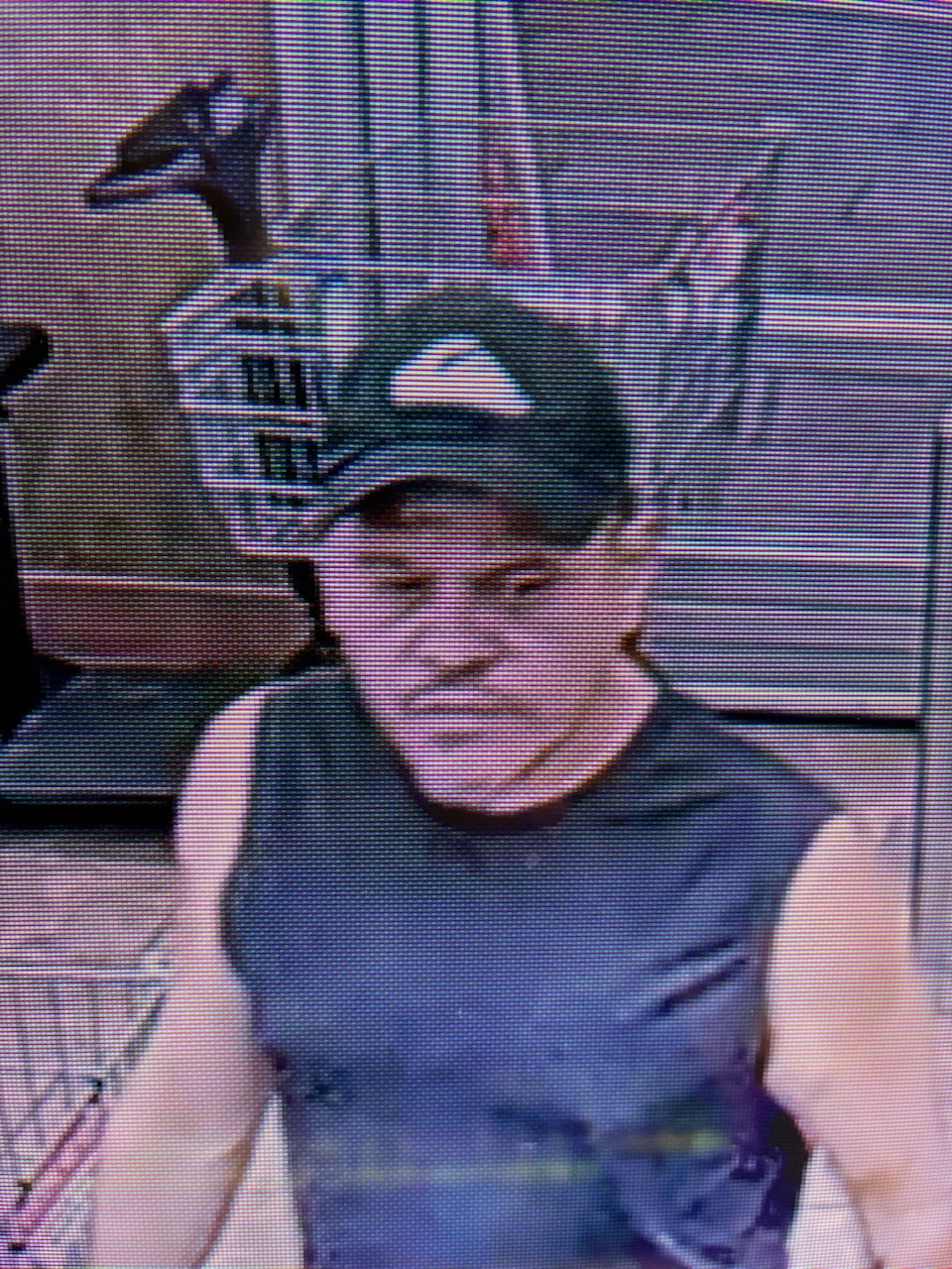 A surveillance-style photo of a man wearing a dark sleeveless shirt and a baseball cap inside what appears to be a retail store. No clear, readable text is visible in the image.