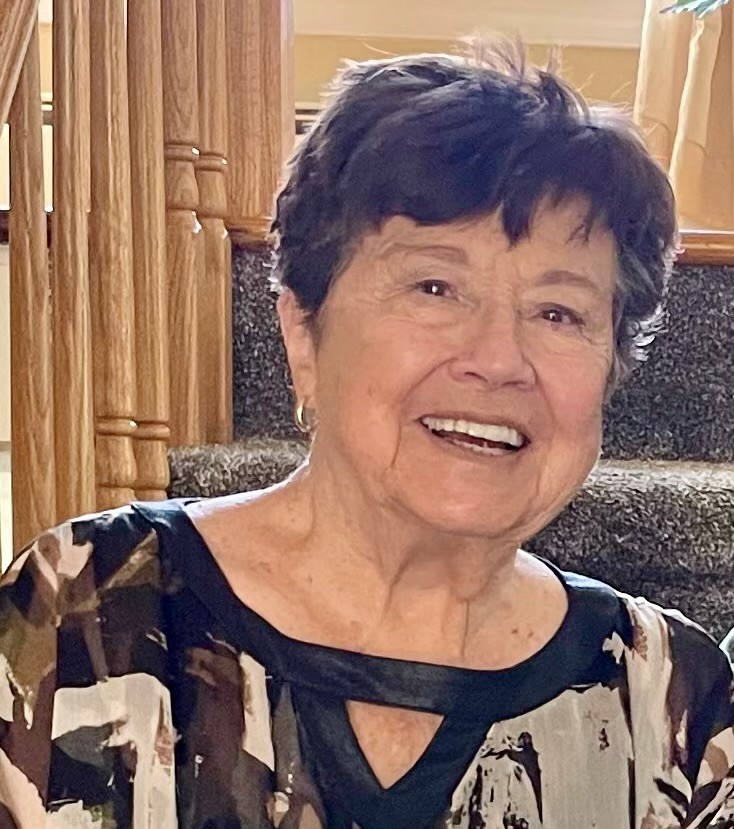 A close-up portrait photo of an older woman smiling indoors near a staircase; there is no visible text to transcribe.