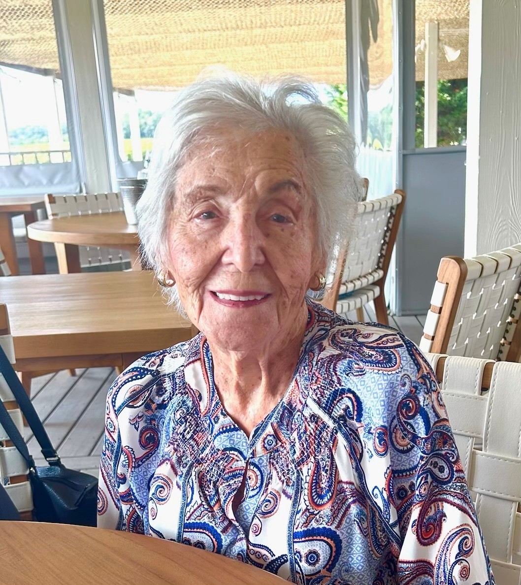 A portrait-style photo of an elderly woman smiling while seated at a table in a bright indoor/outdoor dining area. No readable text is visible in the image.